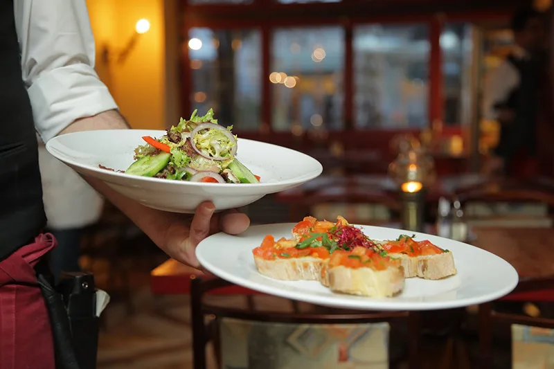 Serveur qui tient deux assiettes d’entrées, une salade et des tartines, dans le restaurant La Table de Trois à Bordeaux